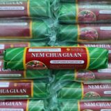 Các thanh Nem Chua Gia An được gói bằng lá chuối xanh mướt, buộc dây chỉ đỏ truyền thống. Bên ngoài là bao bì trong suốt, giúp nhìn rõ từng chiếc nem hồng tươi, điểm xuyết những thớ tỏi trắng và lát ớt đỏ mỏng. Tất cả được sắp xếp gọn gàng, tạo cảm giác tươi mới – thủ công – an toàn vệ sinh. Nhãn dán nổi bật logo Gia An cùng cam kết không chất bảo quản, tôn vinh hương vị quê nhà nguyên bản.